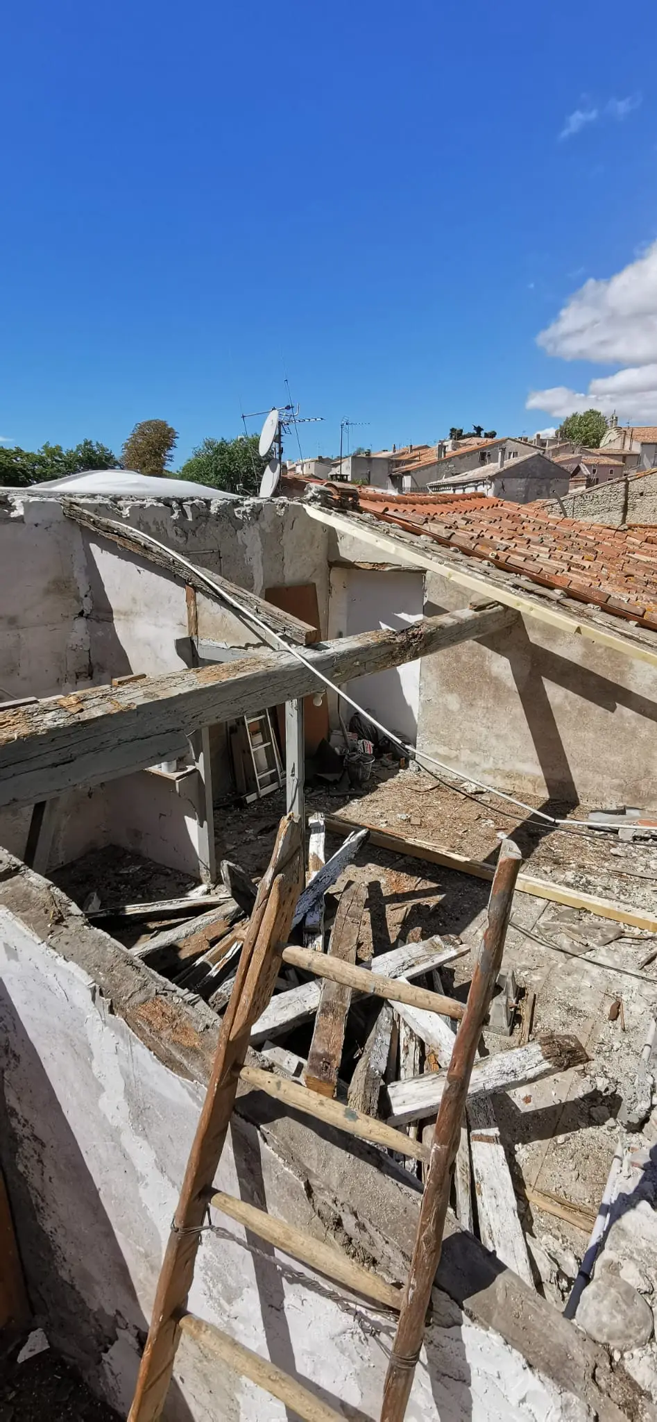 Bâtiment ancien à ciel ouvert : charpente entièrement déposée, murs de pierre mis à nu, échelle de bois appuyée contre l'ouvrage, toits du village au fond