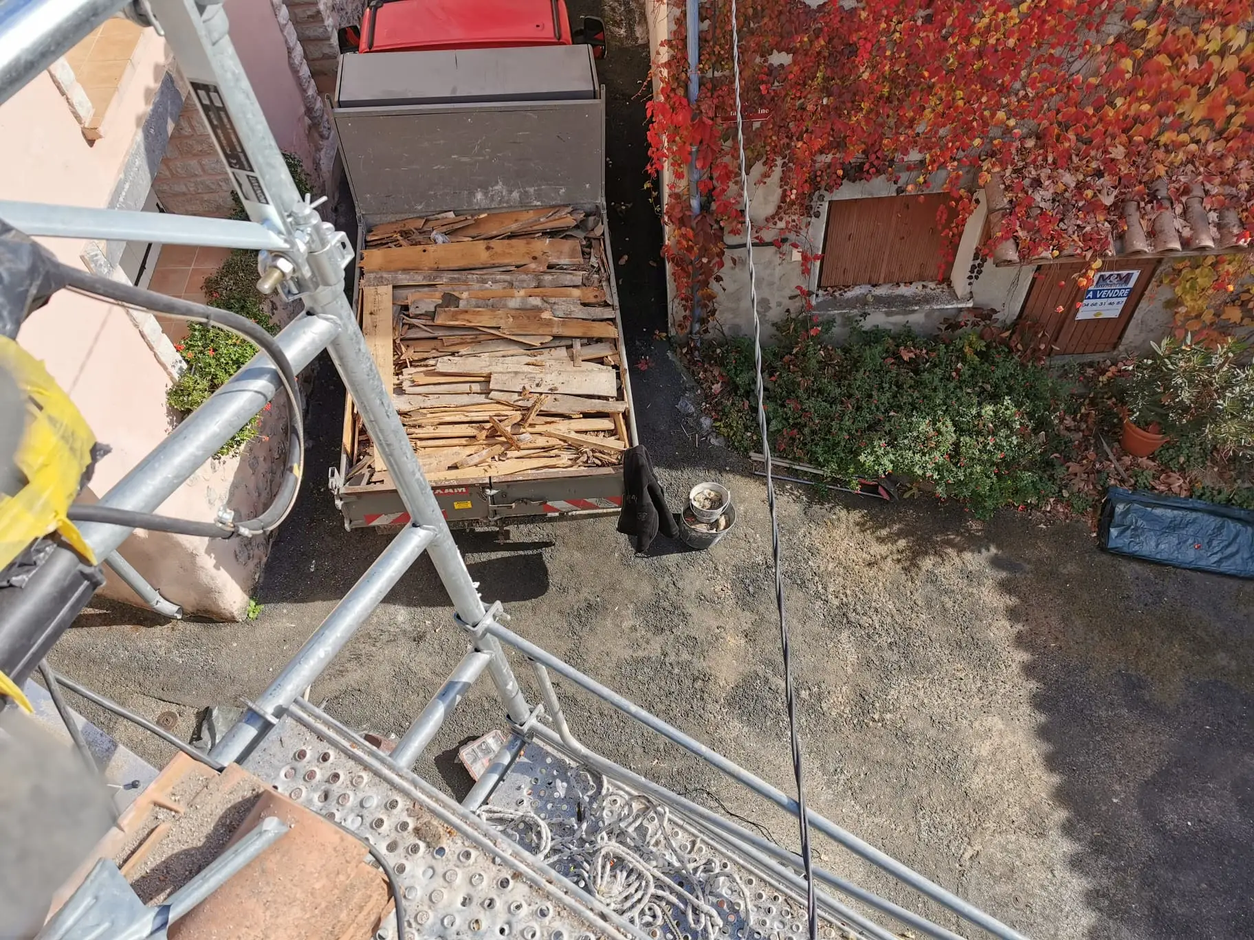 Vue plongeante depuis l'échafaudage sur un camion benne chargé de vieux bois de charpente déposé, le long d'une façade couverte de vigne vierge rouge