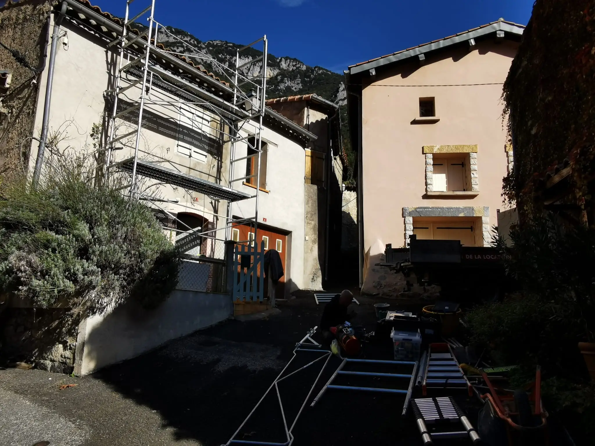 Arriving on a village site: scaffolding erected along a house, equipment and ladders gathered on the ground, with a limestone cliff and blue sky behind
