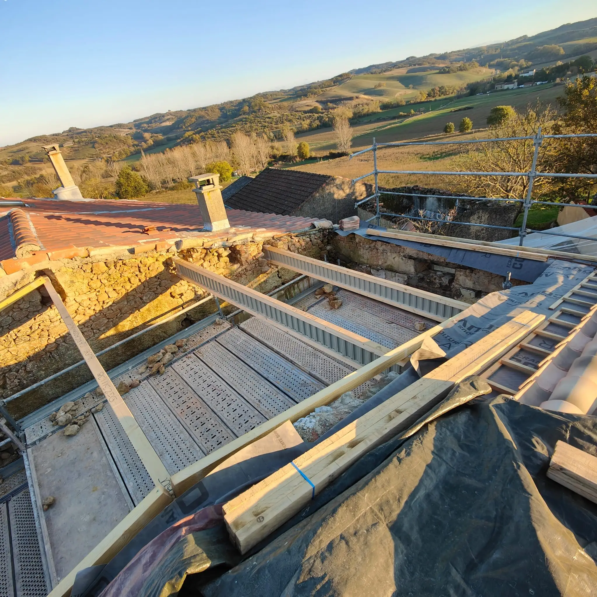 Chevrons neufs posés sur un mur de pierre ancien, vus depuis la toiture en travaux, panorama de collines en lumière rasante