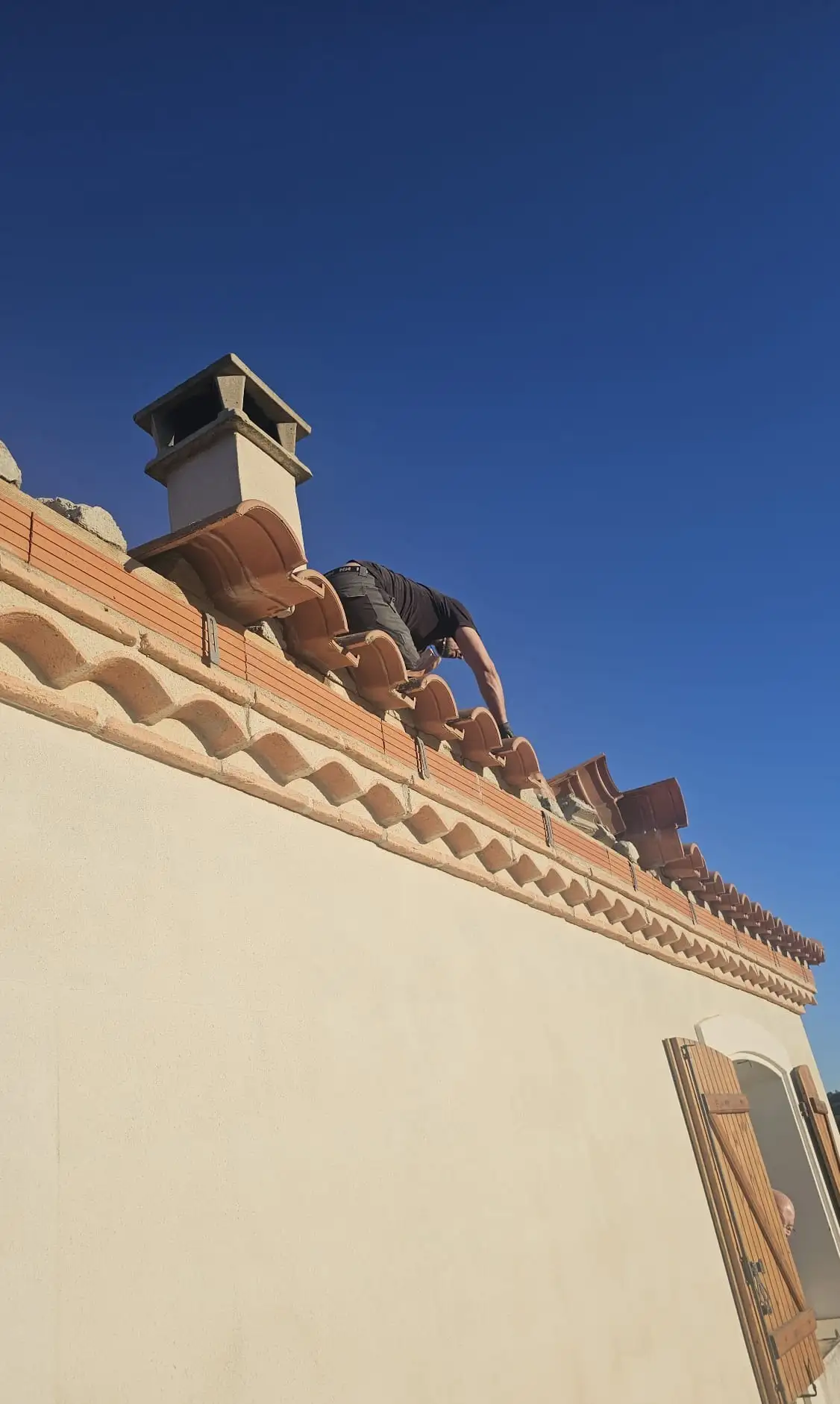 Le couvreur, en contre-plongée sur un ciel bleu profond, pose les tuiles de rive au niveau du dernier rang, au droit d'une souche de cheminée à mitre