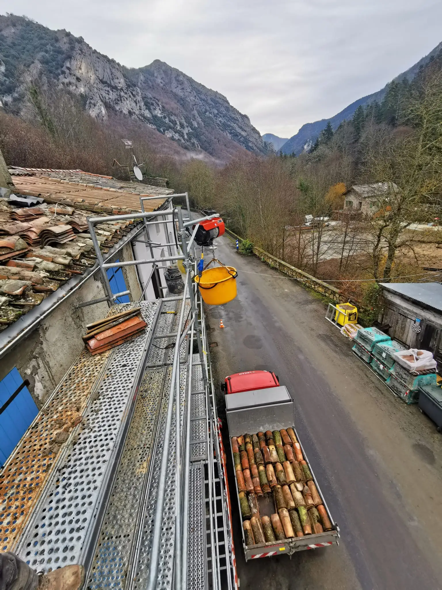 Vue depuis l'échafaudage pendant la dépose : camion benne chargé de tuiles anciennes déposées et treuil de chantier au-dessus de la rue