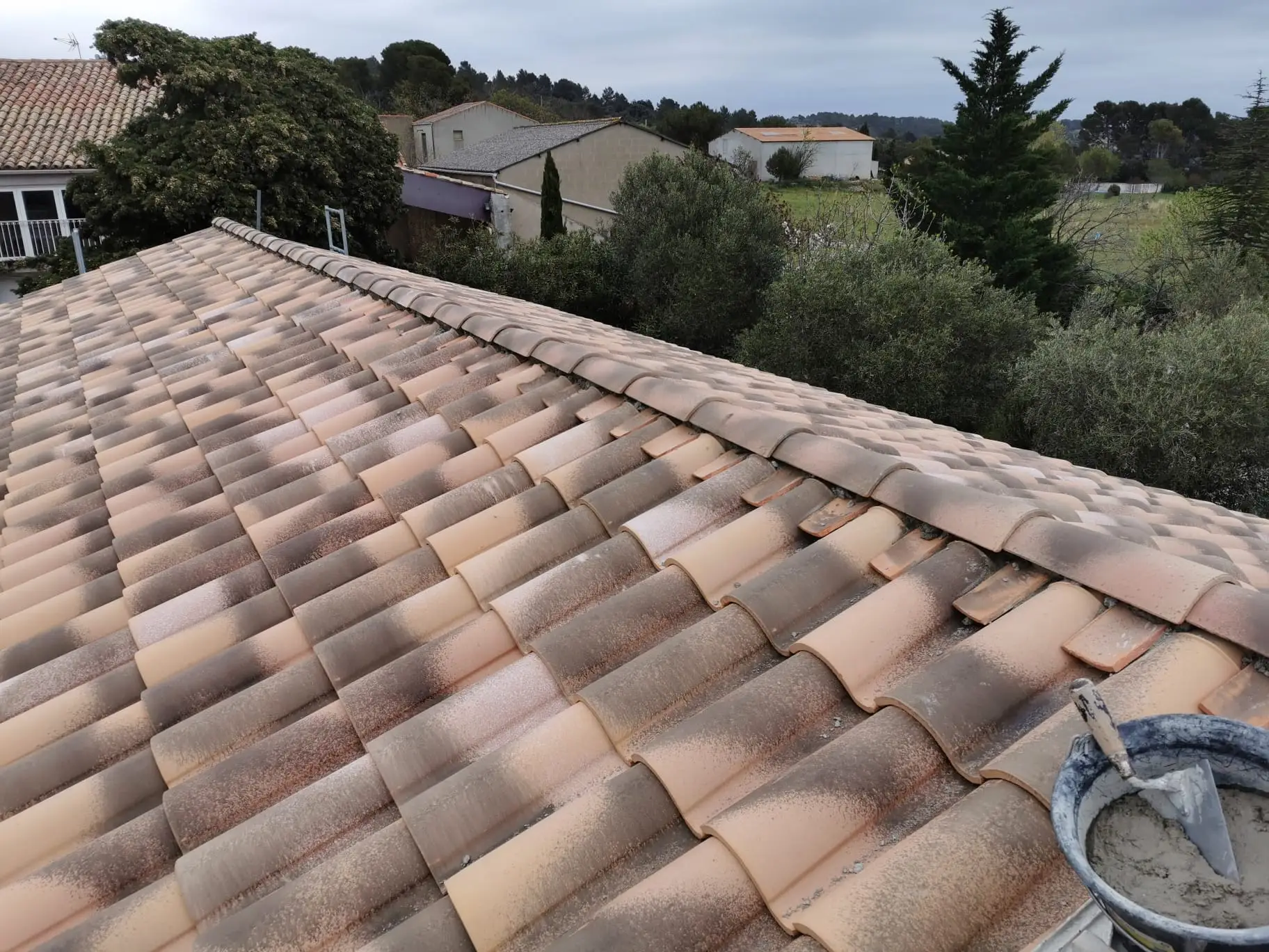 Faîtage scellé au mortier en cours de pose sur une couverture neuve en tuiles panachées, seau de mortier et truelle posés au bord du rampant