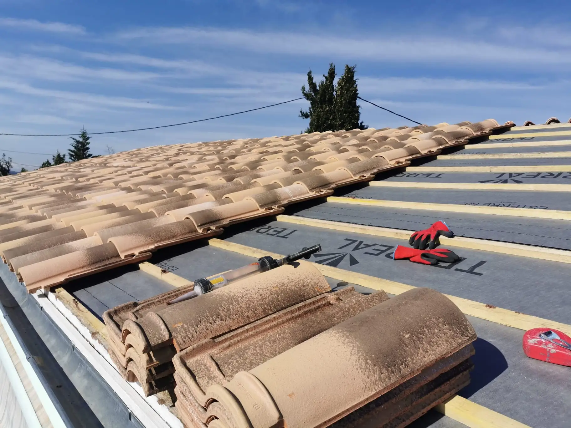 Rive et égout d'une couverture en cours : tuiles neuves posées à gauche, écran de sous-toiture et liteaunage neufs à droite, tuiles de rive et cartouche de mastic au premier plan