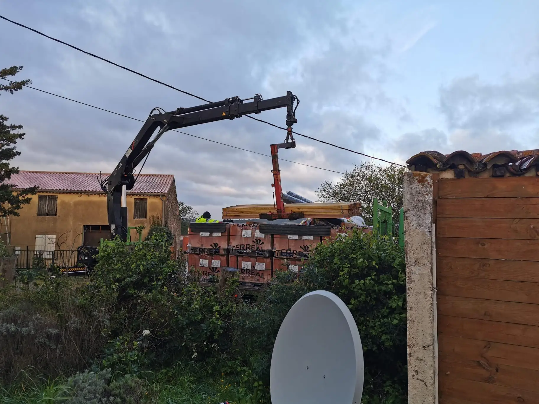Grue auxiliaire de camion déchargeant des palettes de matériaux et du bois près d'une maison de campagne, en fin de journée