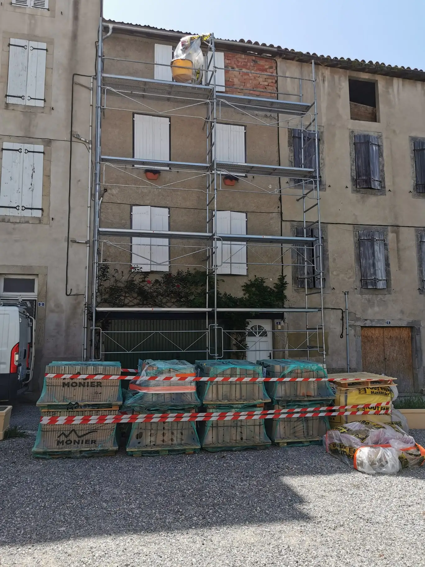 Immeuble de village de trois niveaux sous échafaudage complet, pignon de brique apparente en partie haute, palettes de tuiles et sacs de mortier alignés au pied
