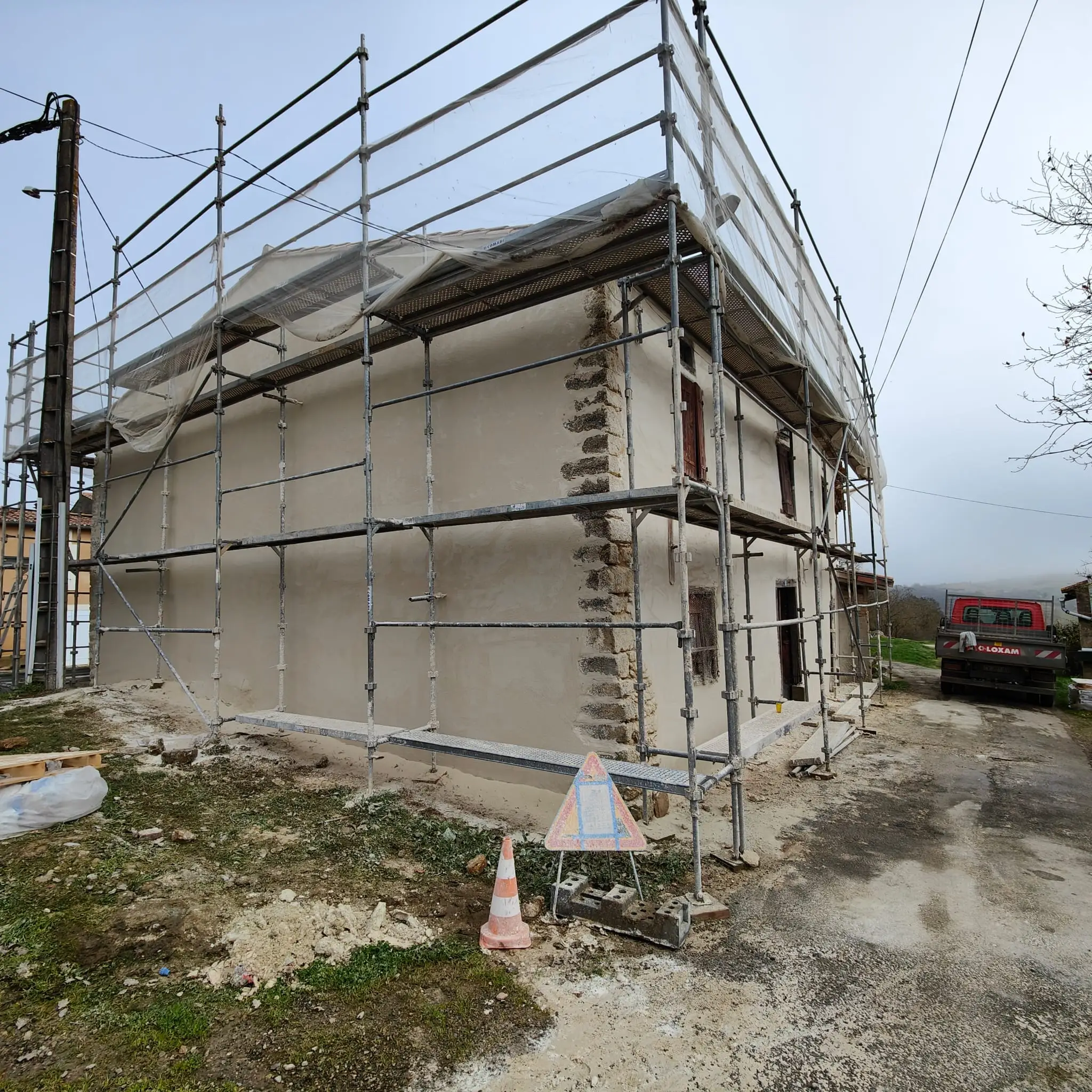 Maison ancienne en cours de rénovation, ceinturée d'un échafaudage sur deux niveaux, chaîne d'angle en pierre laissée apparente et filet de protection en toiture