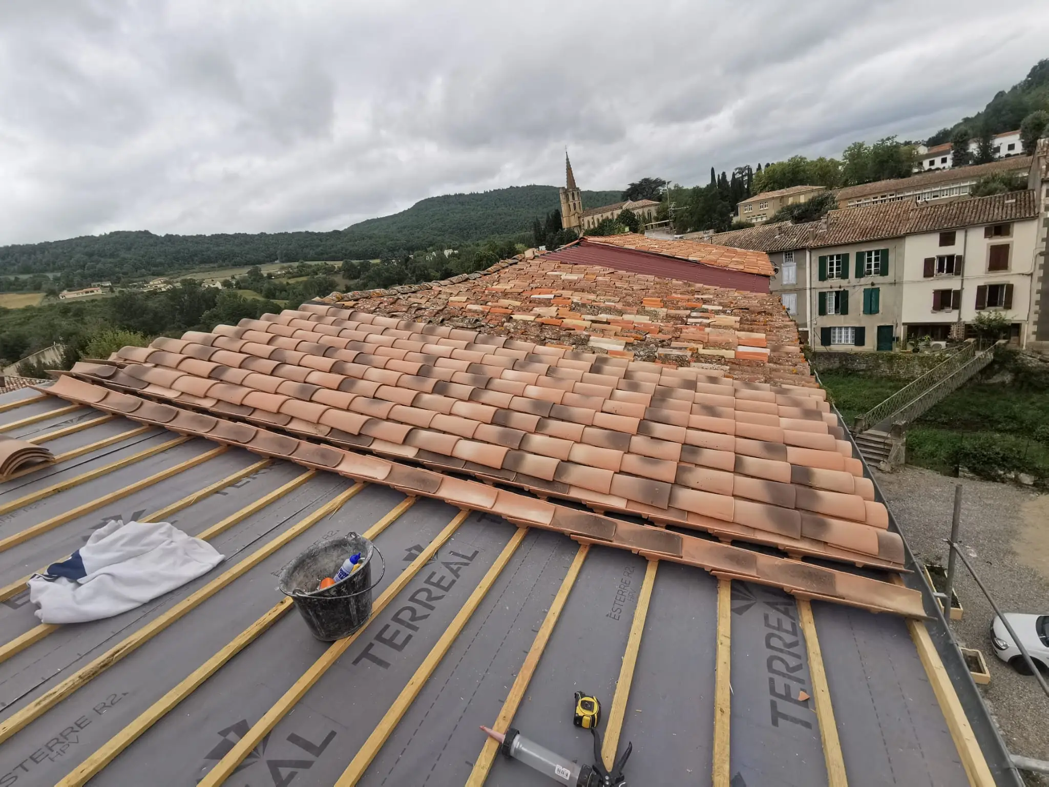 Toiture en cours de réfection vue depuis le faîtage : écran de sous-toiture et liteaux d'un côté, tuiles canal déjà reposées de l'autre, village et collines au fond