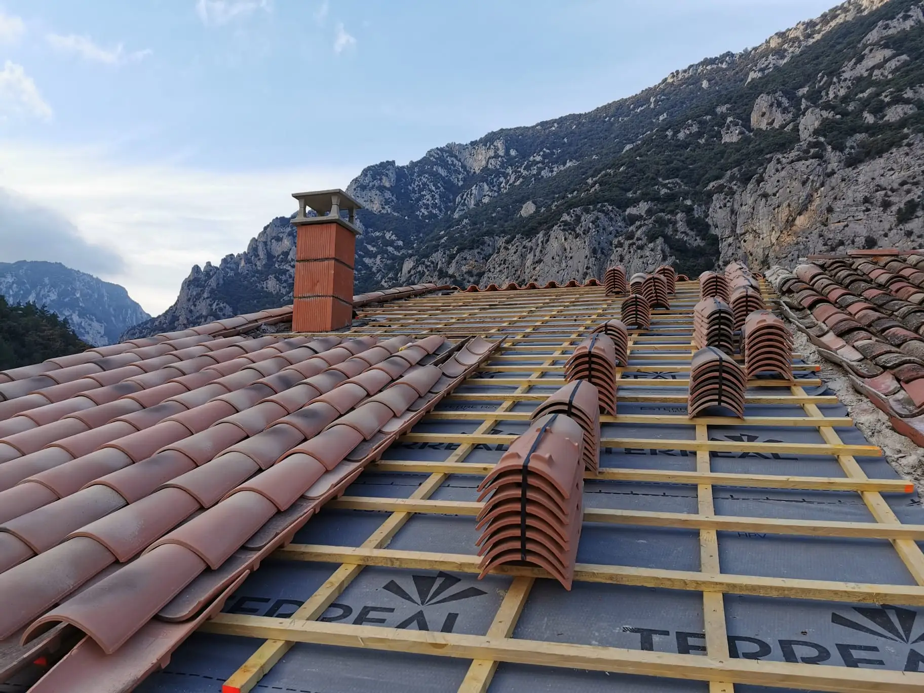 Chantier de réfection : écran de sous-toiture et contre-lattage posés, tuiles canal réparties par rangs sur le rampant avant leur pose, souche de cheminée neuve et falaises en arrière-plan