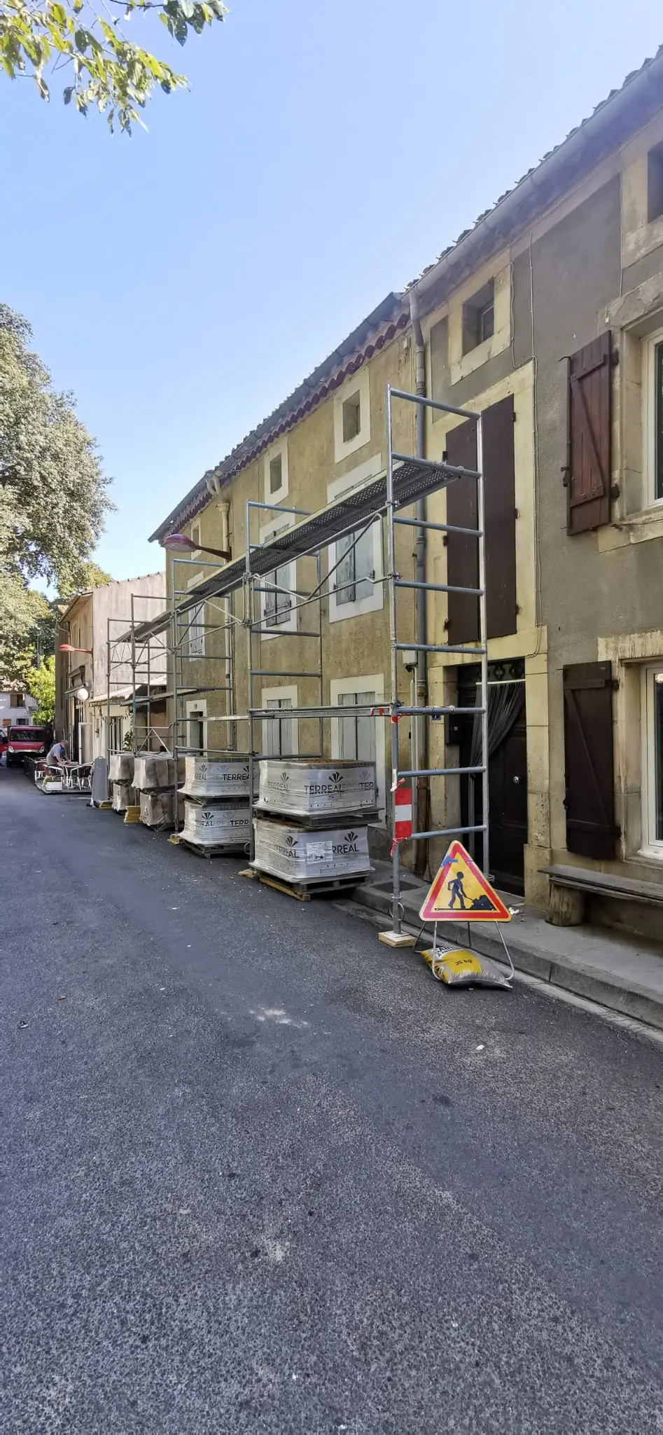 Rue de village aux façades anciennes et volets de bois : échafaudage monté le long d'un bâtiment, palettes de matériaux alignées au sol