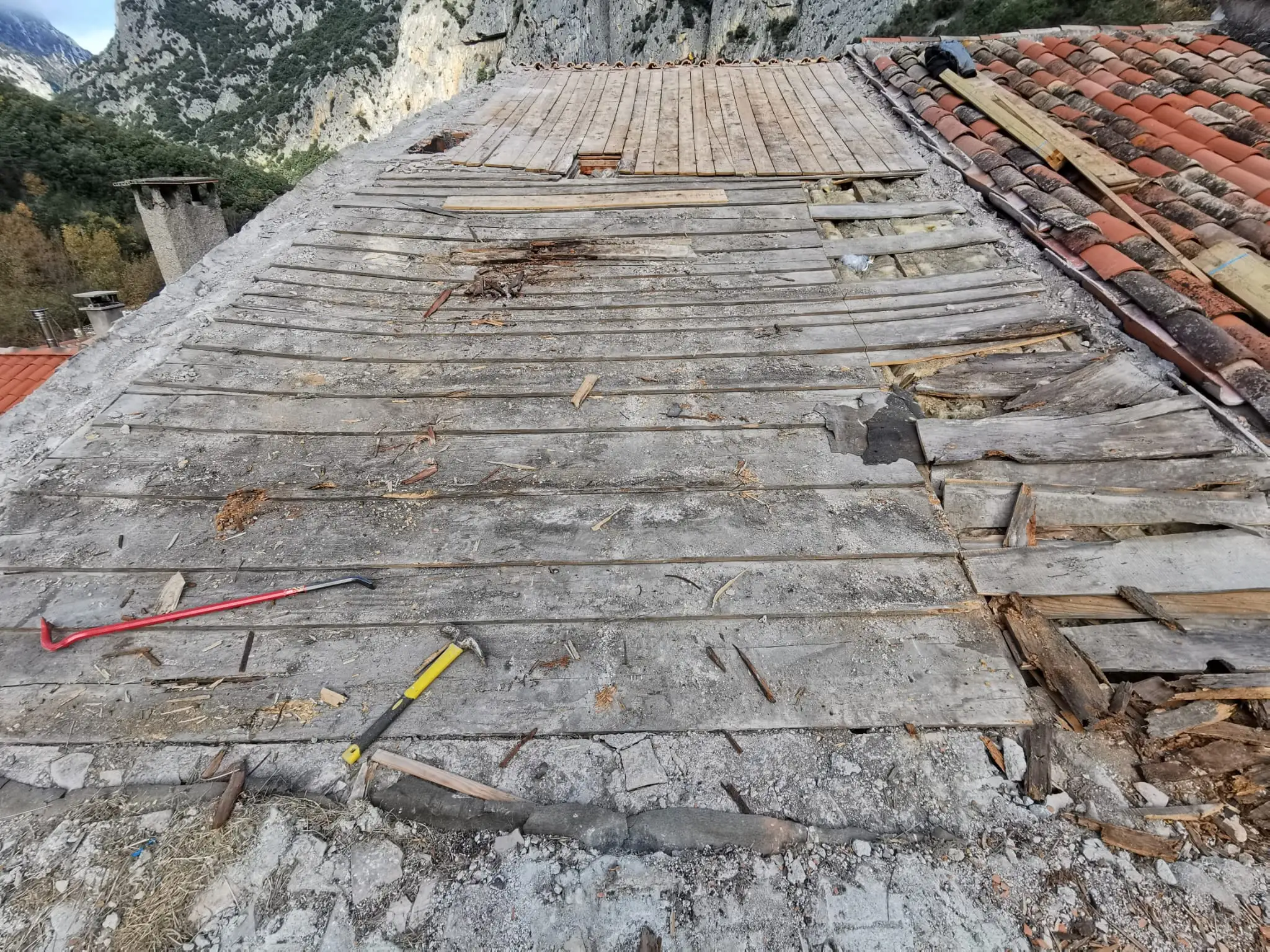Toiture mise à nu : voligeage ancien dégradé et planches manquantes, marteau de couvreur et pied-de-biche posés sur le support