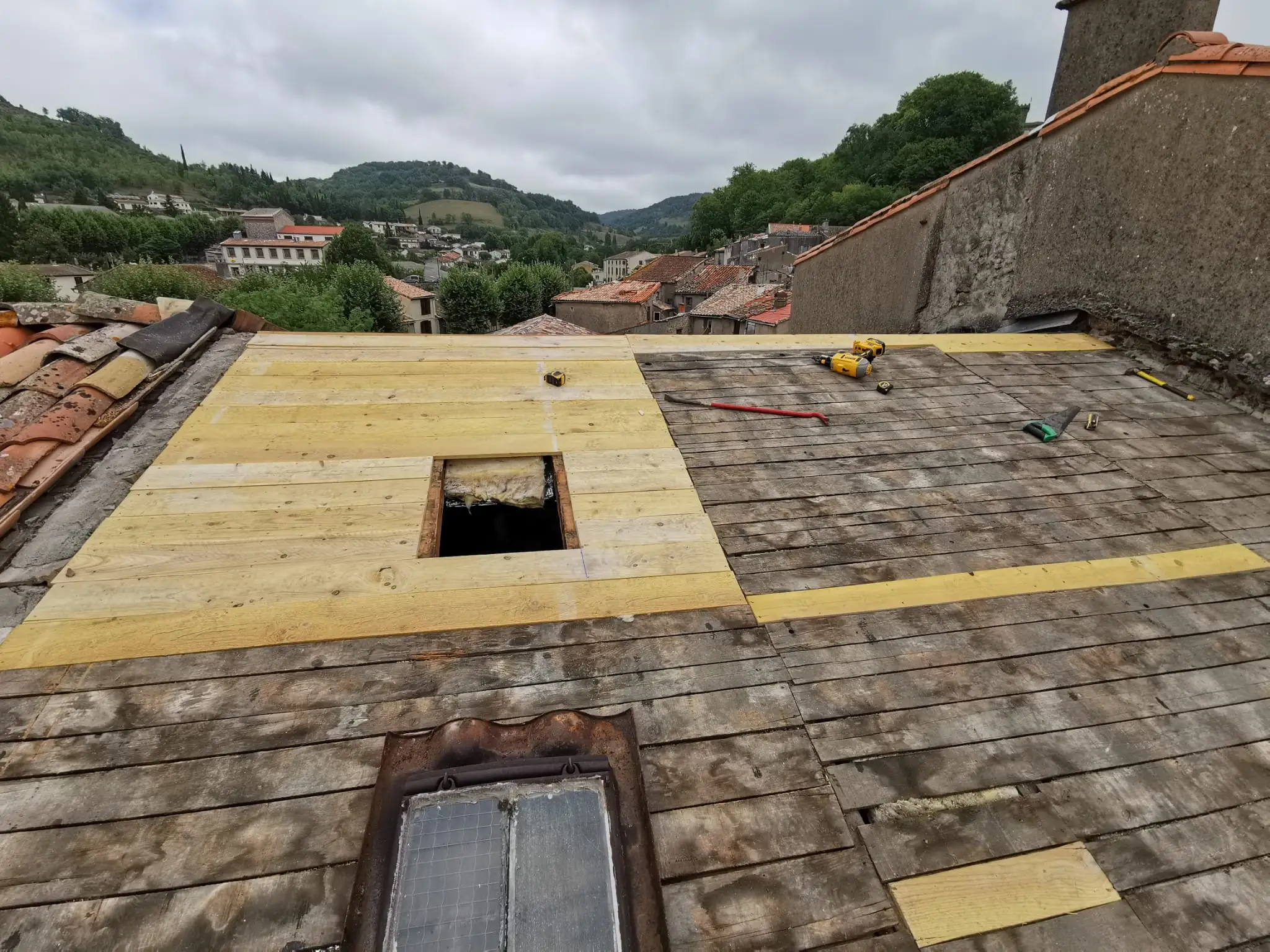 Support de couverture repris : planches anciennes grisées remplacées par endroits par des bois neufs, trémie ouverte, outils posés ; village et collines en arrière-plan